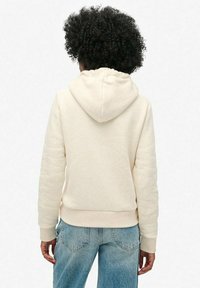Persona con cabello rizado que lleva una sudadera crema y jeans azules, vista desde atrás sobre un fondo blanco liso.