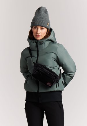 Jonge vrouw die een grijze gebreide muts, groene donsjas, zwarte crossbodytas en zwarte broek draagt, staand tegen een effen achtergrond.