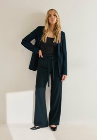 Ciemnozielona marynarka na czarnym topie, zestawiona z szerokimi spodniami i czarnymi butami. Gładki materiał z wiązanym detalem w talii.
