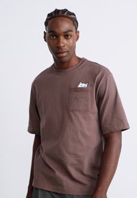 Tânăr cu părul împletit, purtând un tricou maro cu un mic design al unei pisici albe care se arată din buzunarul de la piept, pe un fundal simplu.