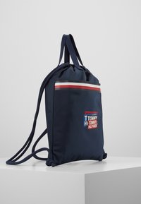 Mochila de cuerda azul marino con rayas rojas, blancas y azules y logo de Tommy Hilfiger, colocada sobre un pedestal blanco contra un fondo liso.