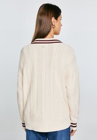 Cremfarbener Strickpullover mit Fischgrätenmuster, gerippter Taille und weinroten Streifenakzenten am Kragen und an den Bündchen.