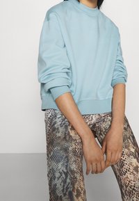 Felpa azzurro chiaro con scollatura arrotondata e spalle scese, abbinata a pantaloni con stampa serpente marroni e beige. Texture liscia.