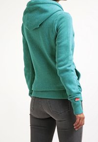 Sudadera color turquesa con tejido texturizado, capucha ajustable con cordón y puños acanalados. Presenta un pequeño logo en la manga izquierda.