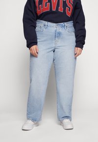 Osoba ubrana w jasnoniebieskie, wysokie dżinsy, białe sneakersy i granatowy sweter z dużym napisem "LEVI'S" na czerwono.