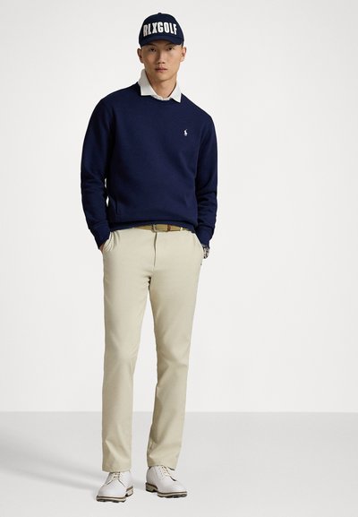 Marine sweatshirt over een witte blouse, beige broek en witte golfschoenen. Een marine pet met de tekst "RLXGOLF" maakt de outfit compleet.