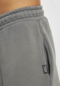 Pantalones deportivos grises con cintura acanalada, bolsillo lateral y una pequeña etiqueta negra con las letras "DEF" en el lado izquierdo. Material suave y texturizado.