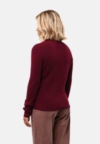 Blondýnka s vlasy po ramena, oblečená v burgundském svetru s dlouhým rukávem a hnědých kordových kalhotách, stojíc í otočená vlevo na bílém pozadí.