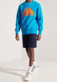 Sudadera turquesa con logo naranja, pantalones cortos marinos y zapatillas blancas. La sudadera tiene mangas largas y puños acanalados. Atuendo casual.