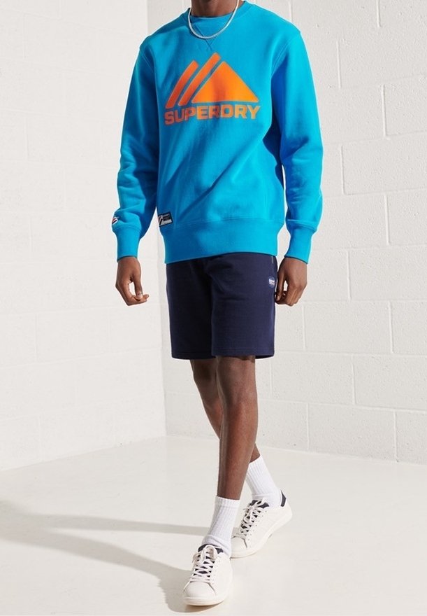 Sudadera turquesa con logo naranja, pantalones cortos marinos y zapatillas blancas. La sudadera tiene mangas largas y puños acanalados. Atuendo casual.