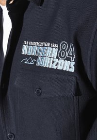 Hemd in Marineblau mit strukturiertem Stoff, zwei Brusttaschen und gestickten Details, darunter "NORTHERN HORIZONS" und Berggrafiken.