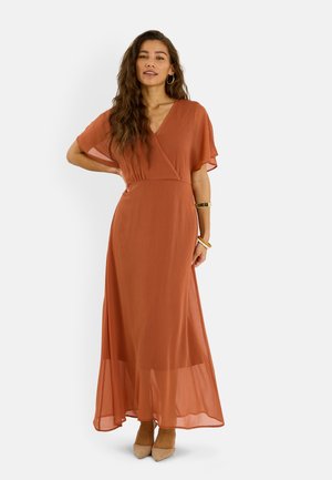 Mujer con cabello largo y ondulado vistiendo un vestido largo cruzado de color naranja óxido y tacones beige, complementada con pulseras y pendientes de aro.