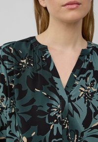 Blumenbluse in Dunkelgrün mit schwarzen und beige Mustern, mit einem V-Ausschnitt und weichem, fließendem Stoff mit gerafften Details.