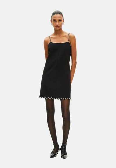 Robe noire sans manches avec ourlet festonné et bordure contrastante, en tissu lisse ; le mannequin porte des collants noirs et des chaussures à talons brillantes.