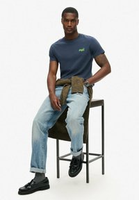 T-shirt navy a maniche corte con logo verde, abbinato a jeans strappati blu chiaro e mocassini neri. Un maglione marrone è annodato in vita.