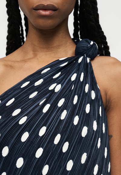 Γυναίκα που φοράει μπλε σκούρο πλισέ τοπ με έναν ώμο και μεγάλα λευκά πουά, στερεωμένο με υφασμάτινο κόμπο στον ώμο.