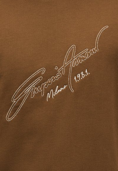 Καφέ ύφασμα με λευκό ραμμένο κείμενο που γράφει «Emporio Armani Milano 1981» με γραφή με καλλιγραφικό στυλ.