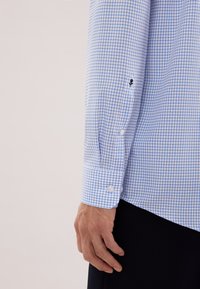 Koszula w niebiesko-białą kratę z mankietem na guziki, z małym czarnym detalem w pobliżu mankietu. Gładka faktura tkaniny.