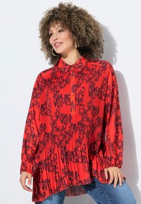 Blusa rossa plissettata con stampa grafica nera, colletto con bottoni, maniche lunghe e polsini elasticizzati. Indossata su jeans blu.