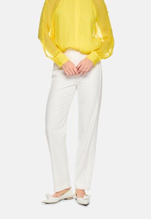 Woman wearing a yellow sheer long-sleeve blouse, white straight-leg pants, and white pointed flats with bow details.