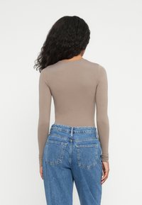 Kvinne med langt, krøllete svart hår iført en tettsittende beige langermet topp og høyhårede blå jeans, vist fra ryggen mot en hvit bakgrunn.