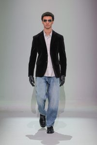 Zwarte fluwelen blazer, wit overhemd met knopen, ruime blauwe jeans, zwarte handschoenen, zwarte schoenen, zonnebril; model dat loopt tegen een neutrale achtergrond.