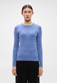 Gaiši zils džemperis ar pīnījuma rakstu, apaļu kakla izgriezumu, fakturētu rakstu, ribota auduma aprocēm un apakšmalai, kā arī nelielu emblēmu uz krūtīm.