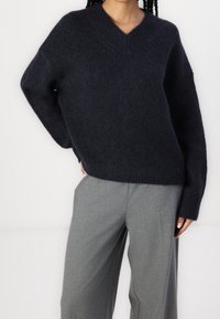 Personne portant un pull en maille duveteuse bleu marine à col en V et un pantalon ample gris, sur un fond blanc uni.
