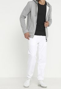 Sudadera gris con cremallera y cordones, combinada con una camiseta negra y pantalones de chándal blancos. Los accesorios incluyen zapatillas deportivas blancas.