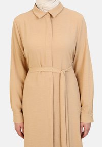 Vestido de manga larga y botón en tela de color beige claro, que incluye un cinturón en la cintura, cuello y puños con detalles de botones. Textura suave.
