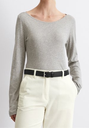 Henkilö, jolla on yllään vaaleanharmaa pitkähiha-paita, joka on tungettu korkeavyötäröisiin valkoisiin puuvillahousuihin, musta vyö, käsi taskussa.