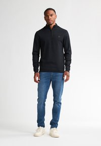 Pullover nero con zip e finitura testurizzata, dotato di un piccolo logo sul petto, abbinato a jeans blu e sneakers bianche.