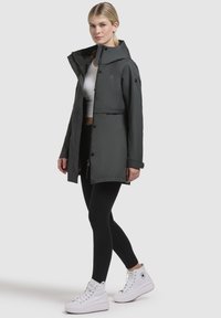 Donkergrijze waterdichte jas met capuchon, voor knopen en elastische manchetten. Gepaarde met zwarte legging en witte hoge sneakers.