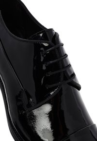 Chaussure habillée en cuir verni noir avec laçage fermé et une fine couture sur fond blanc.