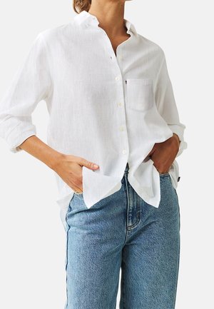 Osoba ubrana w luźną białą koszulę na guziki z podwiniętymi rękawami i niebieskie dżinsy, mająca ręce swobodnie w kieszeniach, na jednolitym tle.