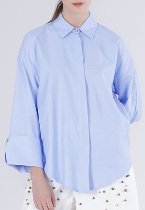 Lyseblå oversized langærmet skjorte med skjulte knapper, båret af en person iført hvide bukser dekoreret med små metalliske nitter.