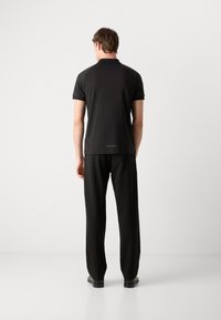 Μαύρη μπλούζα πόλο κοντομάνικη με γιακά, συνδυασμένη με μαύρο παντελόνι. Το ύφασμα φαίνεται λείο, με ελάχιστες λεπτομέρειες.