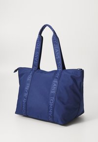 Bolsa de mano azul marino hecha de tejido duradero, con dos asas resistentes y cierre de cremallera. Los detalles incluyen correas texturizadas con la marca "TOMMY JEANS".