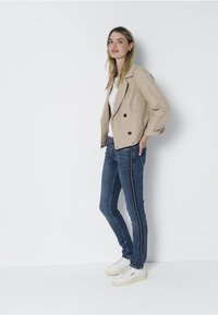 Chaqueta beige corta con detalles de botones, llevada sobre una blusa blanca. Jeans ajustados azul con rayas laterales y zapatillas blancas.