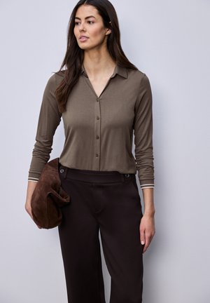 Kobieta ubrana w brązową bluzkę na guziki z długim rękawem i ciemnobrązowe spodnie, trzymająca w lewej ręce złożoną brązową zamszową kurtkę.