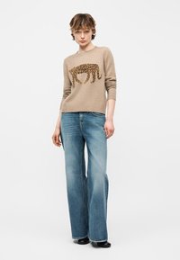 Jeune personne portant un pull beige à imprimé léopard et un jean bleu à jambes larges, debout devant un fond neutre.