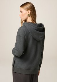 Felpa con cappuccio grigio scuro realizzata in materiale morbido, con cappuccio regolabile, fessure laterali e polsini a costine, indossata su pantaloni grigi.