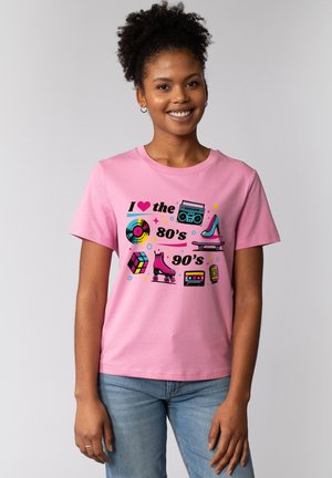 Lächelnde Frau, die ein pinkes T-Shirt mit bunten Symbolen aus den 80er und 90er Jahren trägt: Vinyl-Schallplatte, Ghettoblaster, High Heel, Skateboard, Rollschuh, Zauberwürfel, Kassette und Mikrofon.