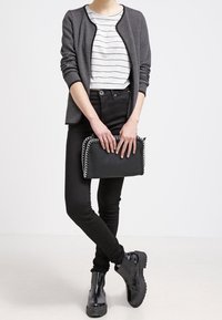 Osoba w czarnych skinny jeansach, czarnych botkach za kostkę, szarym, teksturowanym kardiganie i białej bluzce w paski, trzymająca torebkę kopertową z czarnym łańcuszkiem.