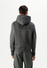 Muž otočený chrbtom, oblečený v tmavosivej mikine s kapucňou, na zadnej strane s veľkým logom a textom Alpha Industries, v kombinácii so zodpovedajúcimi sivými cargo nohavicami.