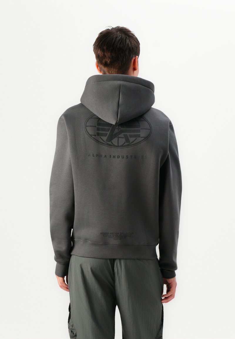 Vyras atsukęs nugarą, vilkintis tamsiai pilką gobtuvą su dideliu "Alpha Industries" logotipu ir tekstu ant nugaros, derinamas su atitinkamomis pilkomis krovinių kelnėmis.