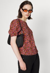 Blusa de manga bufante floral em tecido rosa, laranja e amarelo; mala preta de ombro; óculos de sol laranja; modelo com cabelo curto.