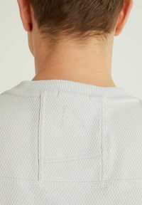 Tył szyi i ramion osoby w białej, teksturowanej koszuli z małym, dyskretnym wyszywanym logo w pobliżu kołnierza.