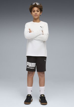 Ragazzo con capelli ricci che indossa una maglia bianca a maniche lunghe, pantaloncini neri PUMA, sneakers nere, calzini bianchi e occhiali da sole sulla testa, in piedi con le braccia incrociate.