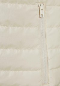 Veste matelassée légère en crème avec surpiqûres horizontales, dotée d'une fermeture éclair blanche avec curseur, texture lisse et sans motifs visibles.